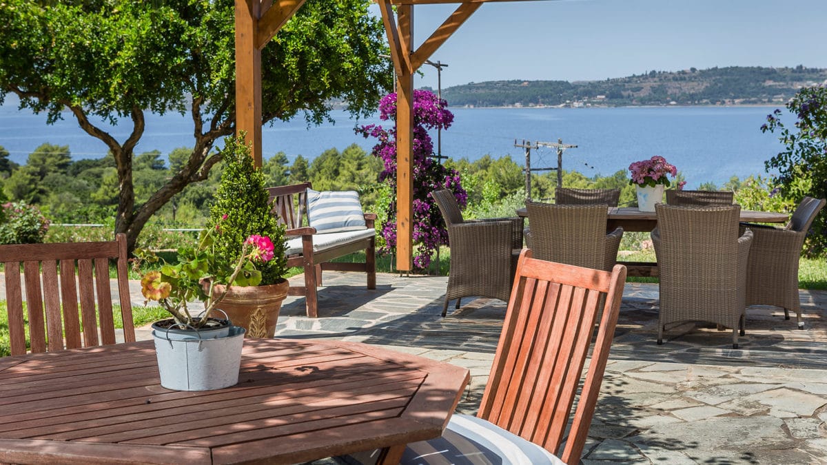 a patio with chairs and a table with plants and a body of water in the background