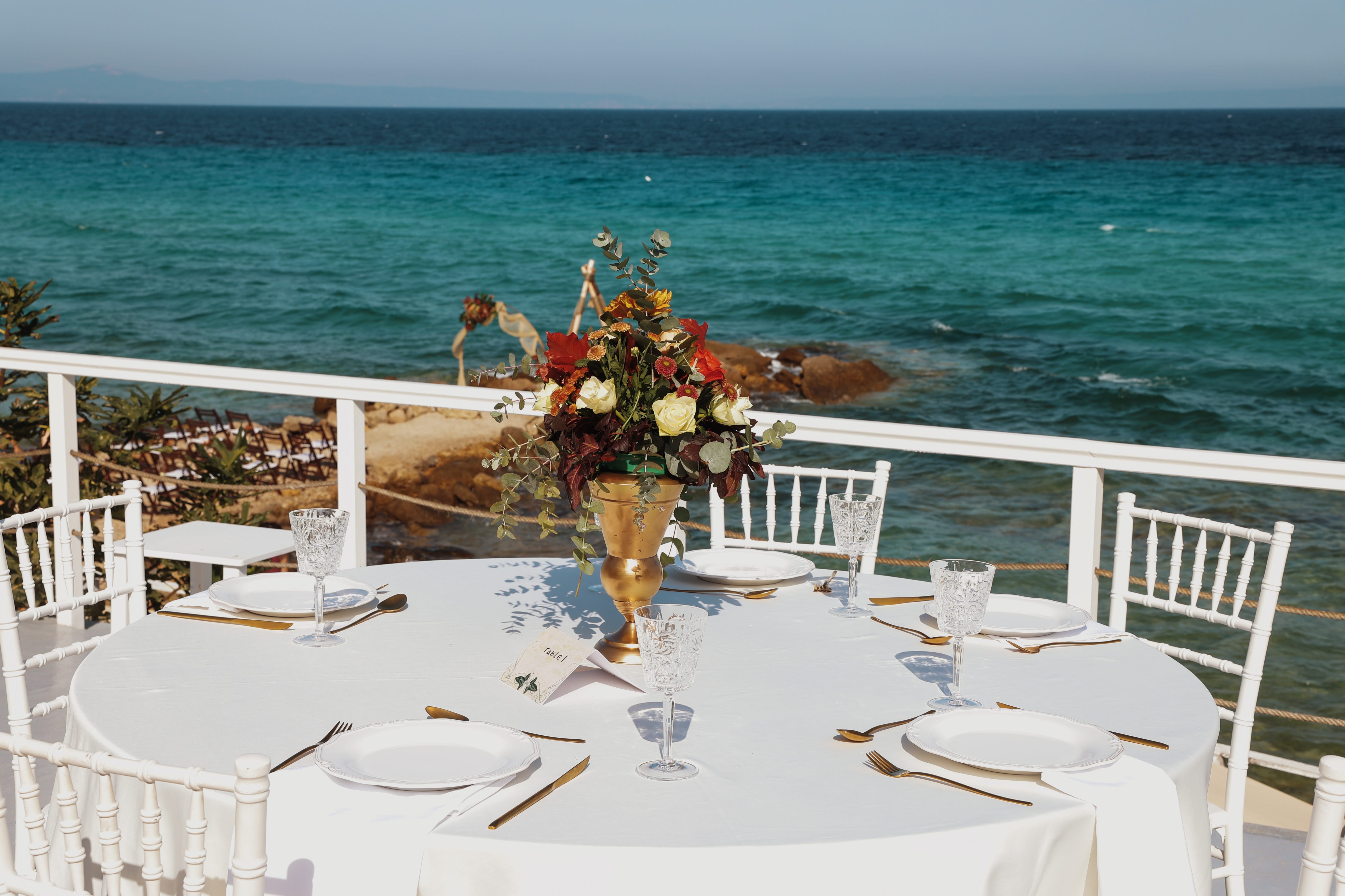 a table with chairs and a vase of flowers on it by the ocean