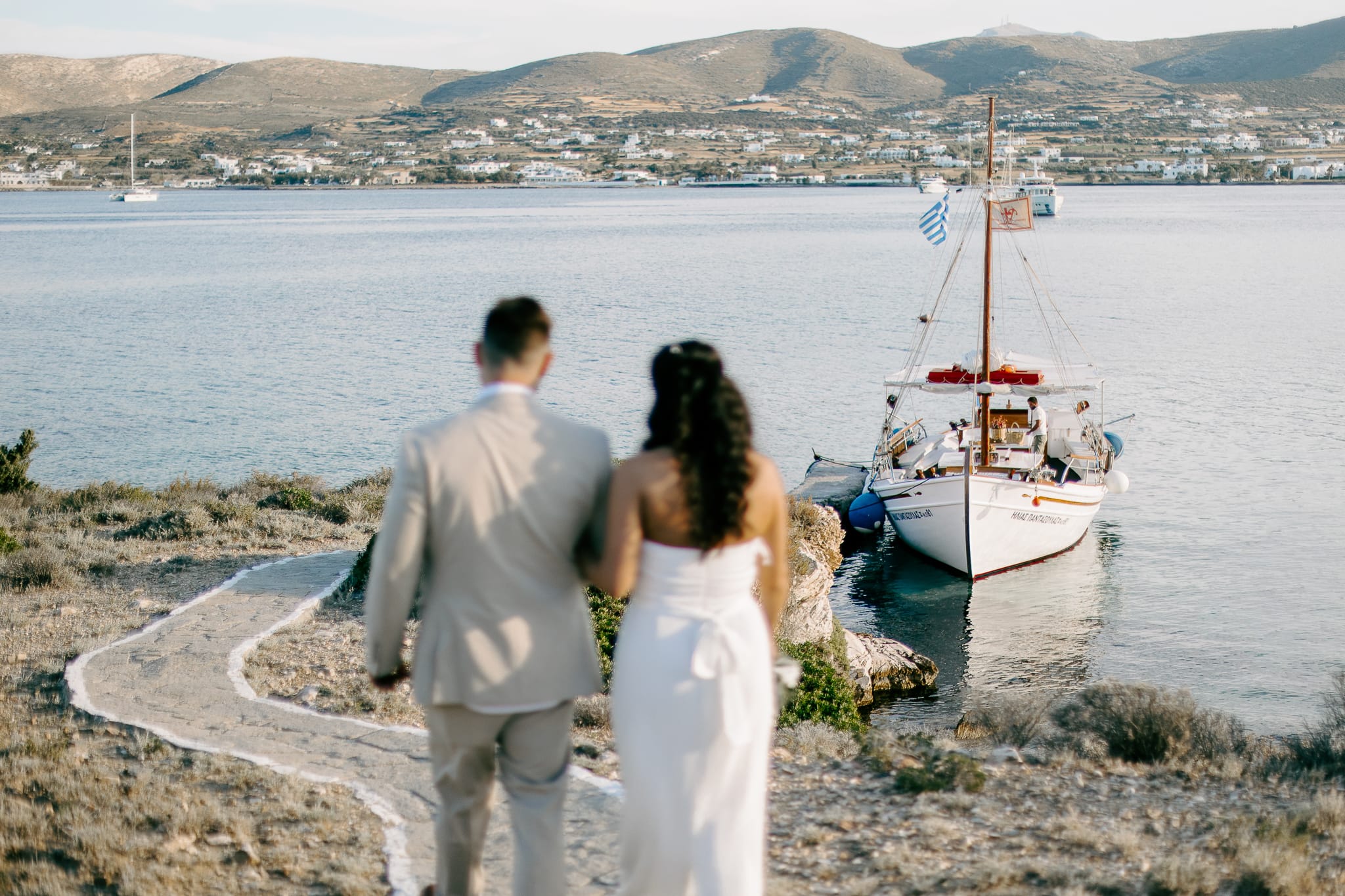 a man and woman standing on a rocky shore looking at a sailboat
