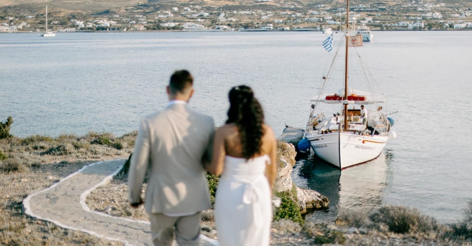 a man and woman standing on a rocky shore looking at a sailboat