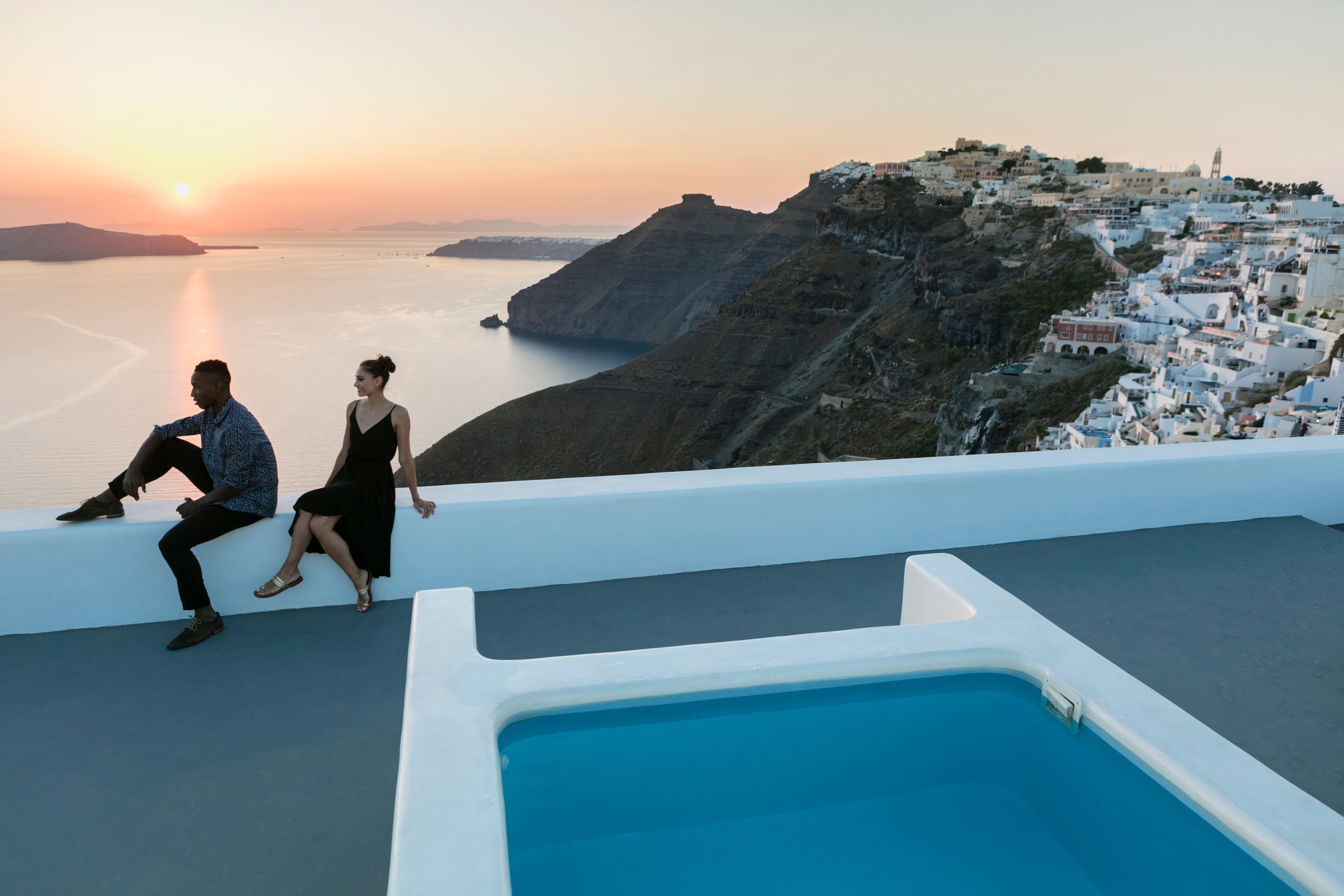 a person and person sitting on a ledge overlooking a body of water with Santorini in the background