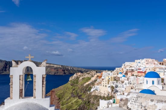 a white building with a bell on top of a hill with a body of water in the background