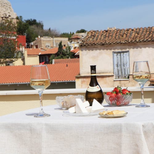 Rooftop dining setup at Kyria Boutique House in Plaka, Athens, featuring a table with wine, cheese, and snacks, overlooking traditional terracotta-roofed houses under a clear blue sky.