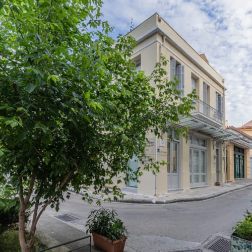Exterior view of Kyria Boutique House with its elegant beige and white facade, located on a peaceful, tree-lined street in the historic Plaka district of Athens, showcasing traditional neighborhood architecture.