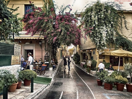 View of a bustling street in Plaka, Athens, featuring pedestrians and outdoor cafes lined with lush pink bougainvillea and white blooms. This vibrant scene captures the essence of a leisurely stroll in the historic heart of Athens, near Kyria Boutique House.