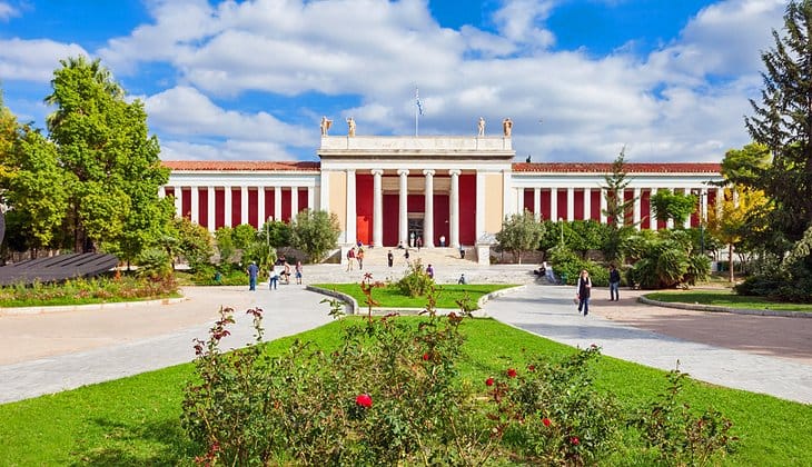 a white building with columns and a lawn in front of it with National Archaeological Museum, Athens in the background