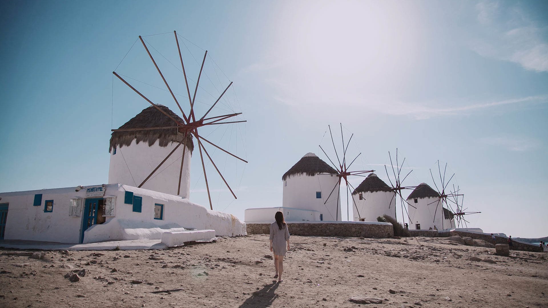 a person standing in front of a windmill on a beach with Mykonos in the background