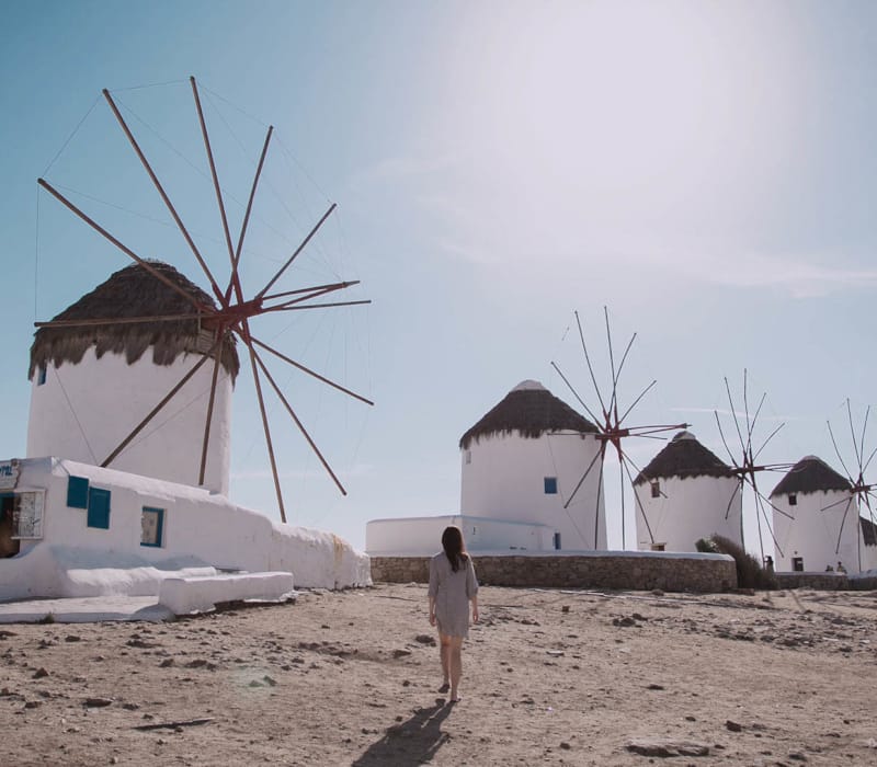 a person standing in front of a windmill on a beach with Mykonos in the background