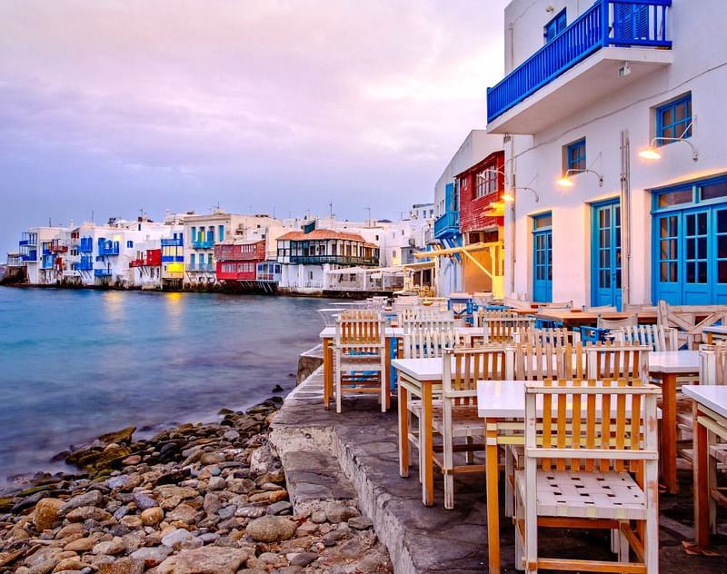 a row of tables and chairs on a rocky beach with buildings in the background