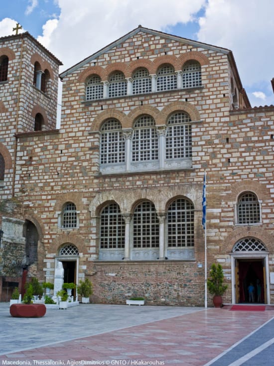 a brick building with a flag in front of it with Hagios Demetrios in the background