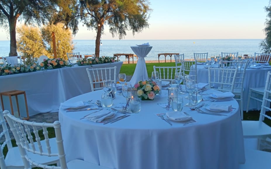 a set table with white chairs and white tables with a view of the ocean and trees