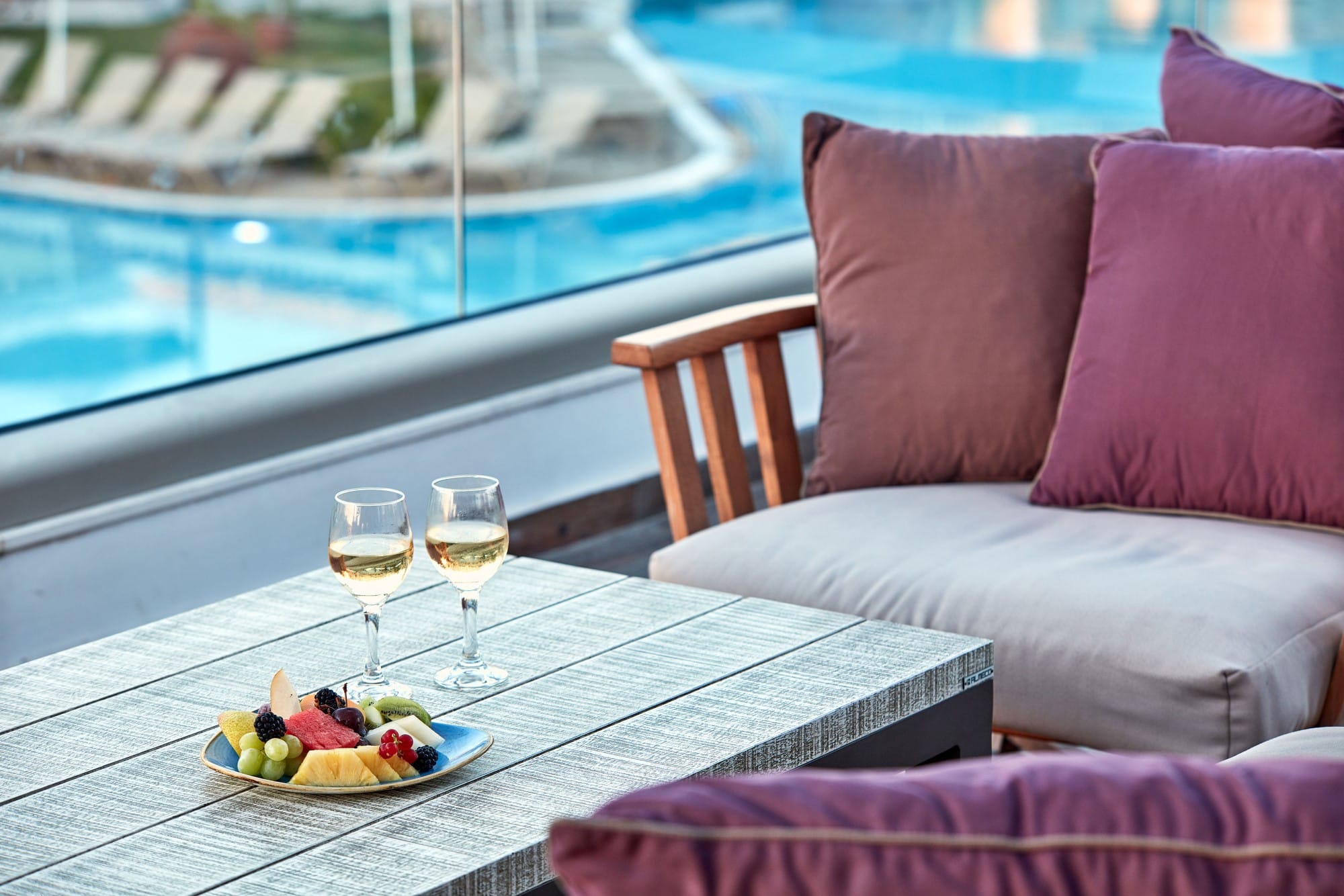 a table with glasses of wine and a plate of fruit on it by a pool