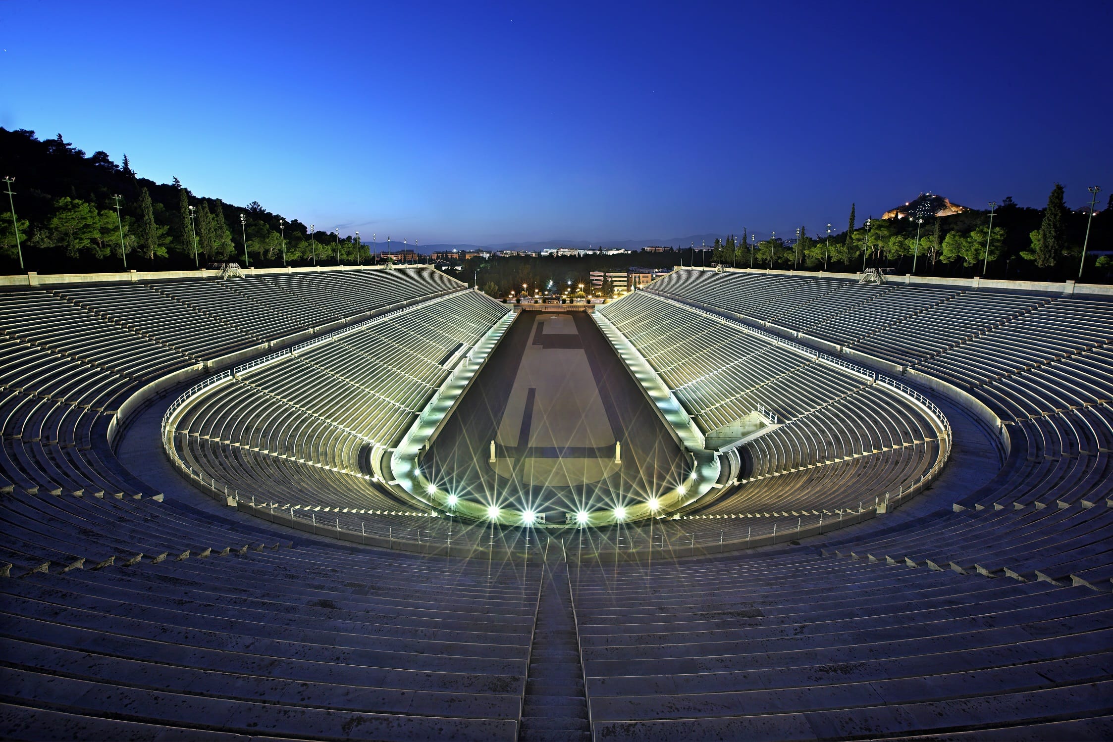 a circular building with lights with Panathenaic Stadium in the background