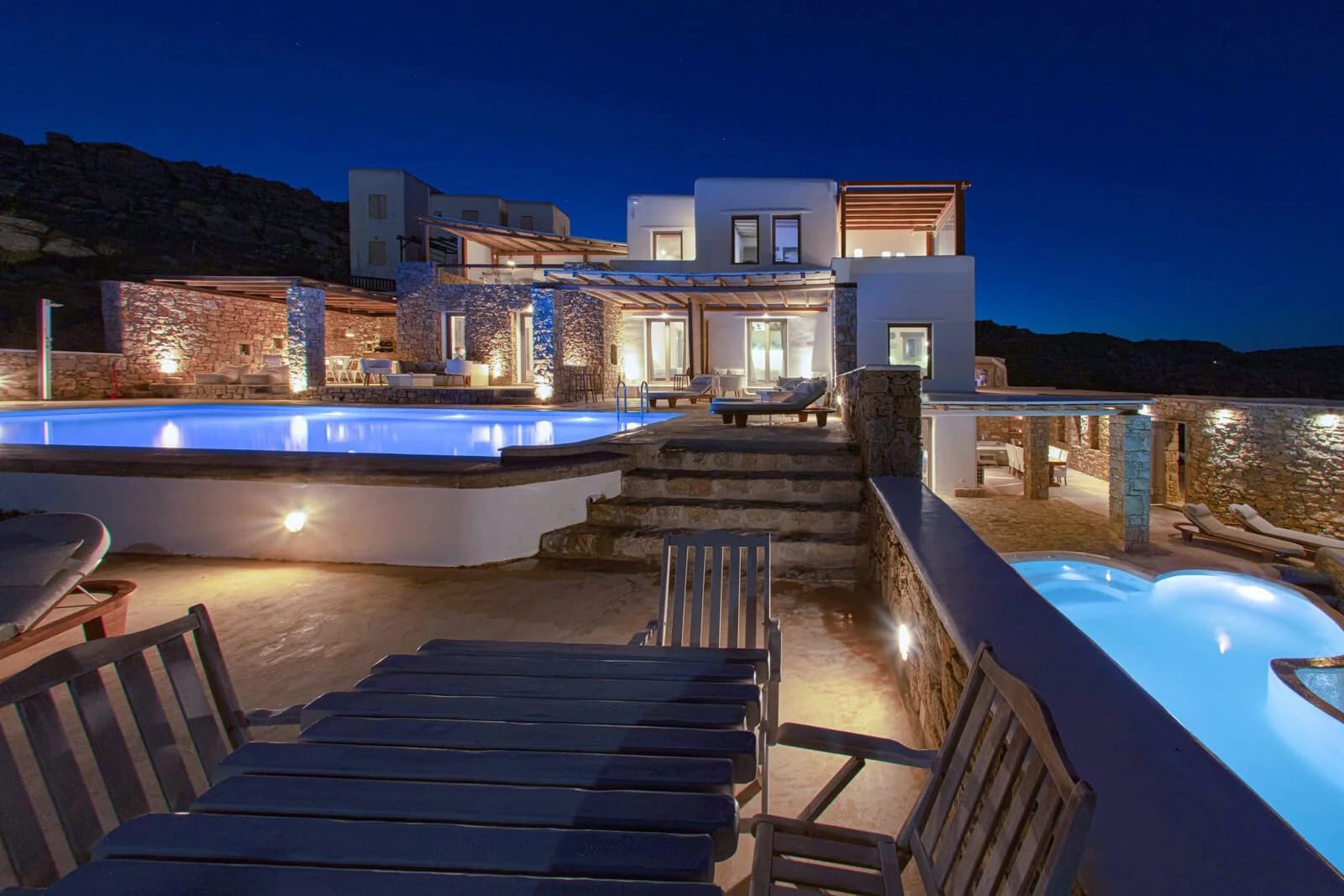 a house with a pool and a deck with chairs and a view of the ocean at night