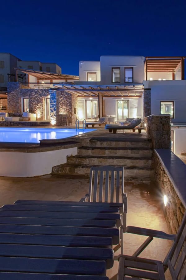 a house with a pool and a deck with chairs and a view of the ocean at night